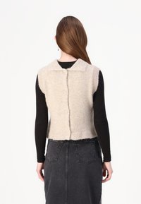 Gilet lavorato a maglia beige con collo a camicia, maniche corte e cinque bottoni lungo la schiena. Indossato sopra una maglia nera a maniche lunghe.