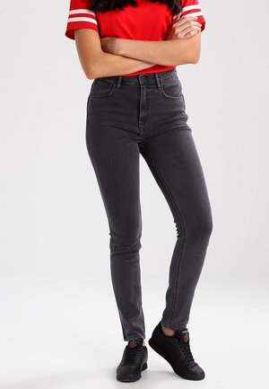 Femme en t-shirt rouge manches courtes avec des rayures blanches, jean skinny gris foncé et baskets noires, debout les bras croisés.