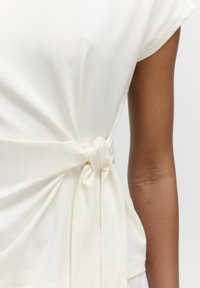 Weiße Bluse aus glattem Stoff mit einer Knoten-Detail an der Taille. Verfügt über kurze Ärmel und leichte Drapierung.