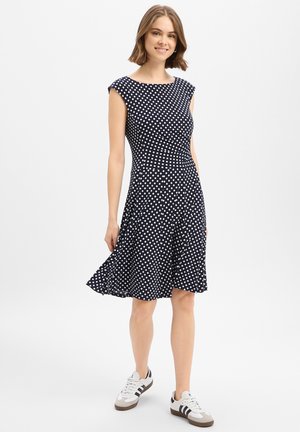 Frau lächelt, trägt ein marineblaues ärmelloses Kleid mit Punkten und weiße Turnschuhe, steht vor weißem Hintergrund.