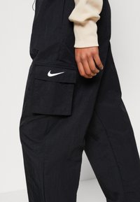 Černé nylonové cargo kalhoty s velkou kapsou se klopou, ozdobené bílým logem Nike. Pomačkaná textura, pohodlný střih.