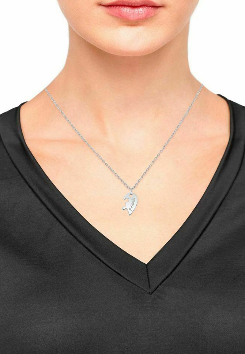 Collana argentata con pendente a forma di pezzo di puzzle inciso con la parola "Amici", indossata da una persona con una maglietta nera a collo a V.