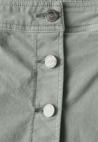 Grüner Cordstoff mit drei silbernen Knöpfen entlang der Vorderseite und strukturierten vertikalen Linien. Die Knöpfe tragen Markenlogos und Text.