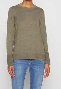Olivegrüner, langärmliger Strickpullover mit gerippten Bündchen und Saum, runder Ausschnitt und subtiler Textur. Getragen mit blauen Jeans.