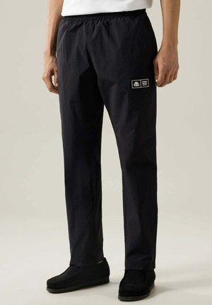 Modello maschile che indossa pantaloni casual neri Kappa con fascia elastica in vita e toppa con logo sulla coscia sinistra, abbinati a scarpe slip-on nere.