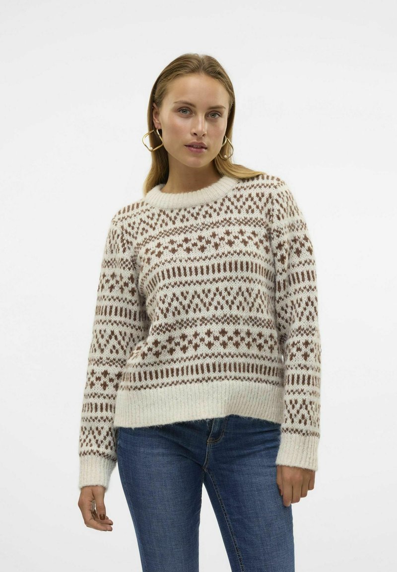Pullover in Creme mit braunen Mustern, gestrickte Textur, runder Hals, und langen Ärmeln. Kombiniert mit blauen Jeans.