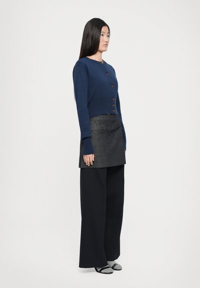 Mornarsko pleteno kratko jopico z lesenimi gumbi, kombinirano z temno denim krilom in širokimi črnimi hlačami. Model stoji v profilu.