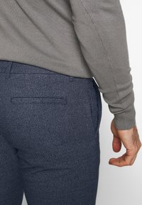 Mörkblå strukturerade byxor med ett bakfick, i kombination med en grå långärmad topp, visar bak- och sidovy.