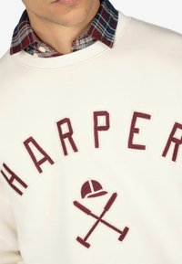 Sudadera blanca con "HARPER" bordado en color burdeos. Cuenta con un cuello de una camisa de cuadro debajo en rojo, azul y blanco.
