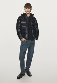 Zwarte pufferjas met een glanzende afwerking, ritssluiting en capuchon, gecombineerd met donkerblauwe jeans en zwarte schoenen. Eenvoudig ontwerp, geen patronen.