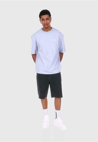 Lyseblå kortermet t-skjorte med en liten hvit logo, kombinert med mørkegrå shorts og hvite sneakers, stående mot en nøytral bakgrunn.