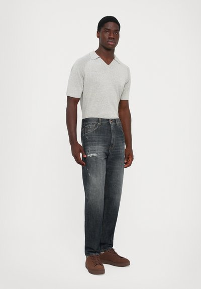 Uomo che indossa una maglietta a maniche corte grigio chiaro in maglia, jeans scuri sbiaditi con strappi e sneakers marroni, in piedi su uno sfondo bianco.