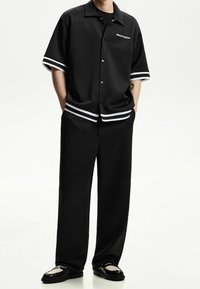 Černá košile s krátkými rukávy a bílými pruhy na manžetách, v kombinaci s širokými černými kalhotami a lesklými mokasíny, představující minimalistický design.