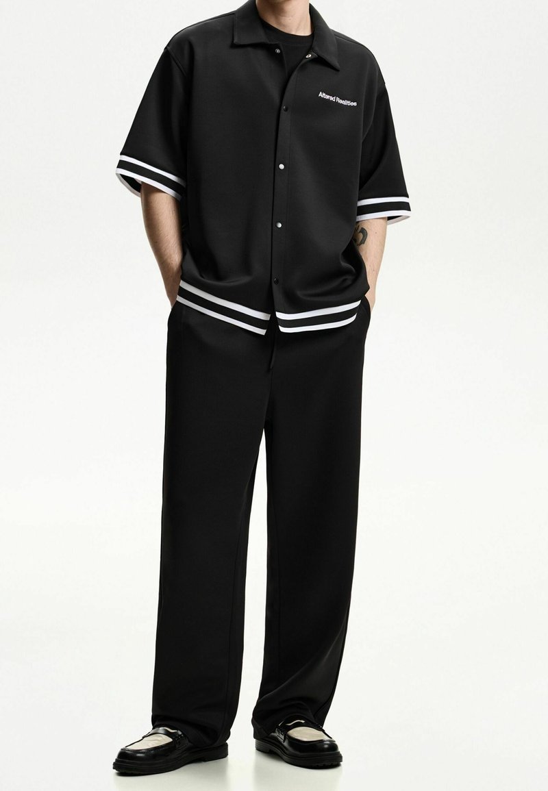 Černá košile s krátkými rukávy a bílými pruhy na manžetách, v kombinaci s širokými černými kalhotami a lesklými mokasíny, představující minimalistický design.