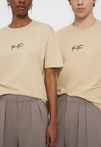 Deux personnes portant des t-shirts beiges avec un texte noir en écriture cursive et des pantalons plissés gris, debout côte à côte sur un fond uni.
