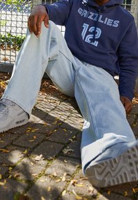 Jeans baggy blu chiaro con una texture sbiadita, abbinati a un hoodie blu navy scuro con la scritta "GRIZZLIES" e il numero "12". Sneakers grigie con suole spesse.