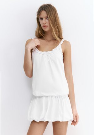 Ung kvinde iført en ærmeløs hvid kjole med flæset halsudskæring og kant, står foran en ensfarvet hvid baggrund.