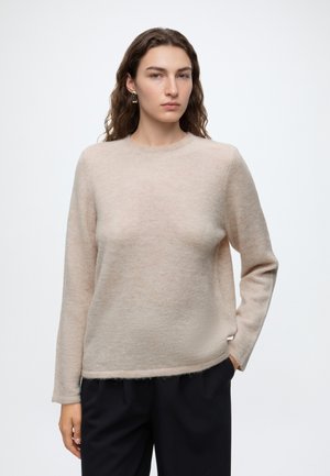Jeune femme aux longs cheveux bruns portant un pull beige et un pantalon noir, debout avec une main dans la poche devant un fond uni.