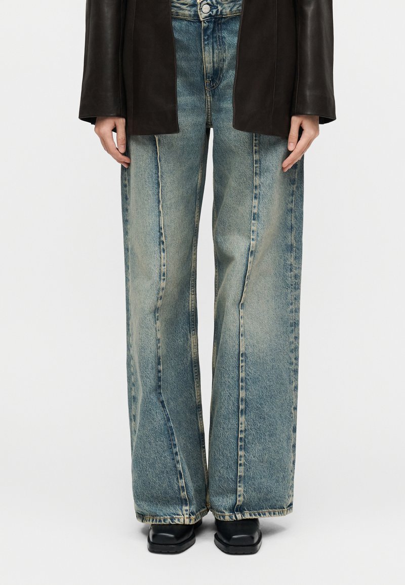 Άτομο που φοράει φαρδιά ξεβαμμένα  μπλε τζιν, μαύρα παπούτσια και σκούρο καφέ μπουφάν, όρθιο με τα χέρια χαλαρά στο πλάι.