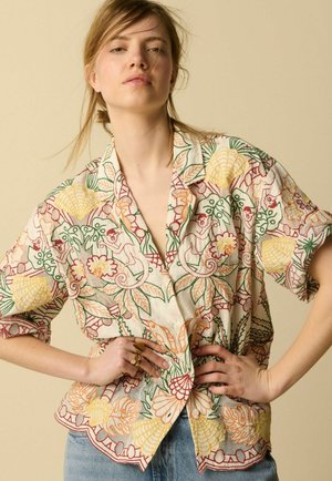 Jeune femme portant une chemise beige ample avec une broderie florale multicolore, debout les mains sur les hanches devant un fond beige uni.