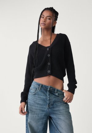 Cardigan nero cropped con lavorazione a maglia testurizzata, scollo a V e chiusura con cinque bottoni. Abbinato a jeans in denim azzurro chiaro a vita alta.