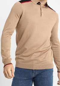 Beige polo tričko s dlouhým rukávem a žebrovanou strukturou, černými a červenými akcenty na ramenou a tříknoflíkovým zapínáním. Jemné logo vyšití na hrudi.