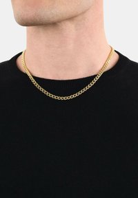 Uomo che indossa una collana d'oro sopra un maglione nero a collo rotondo, collo e mascella visibili su uno sfondo bianco.