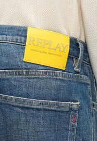 Κοντινό πλάνο μπλε τζιν με κίτρινη ετικέτα της μάρκας Replay και μικρό κόκκινο "R" κεντημένο κοντά στην πίσω τσέπη, φορεμένο με μπεζ πλεκτό τοπ.