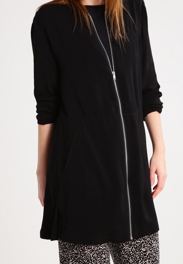 Femme portant une longue veste noire zippée aux manches retroussées et un pantalon noir et blanc à motifs.