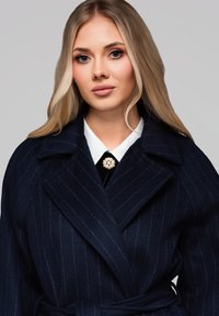 Abrigo envolvente de lana navy con rayas finas, amplios solapas y cintura ceñida. Superpuesto sobre una camisa blanca con cuello y un toque de broche de perlas.
