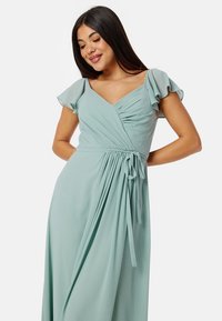 Mintgroene jurk met vloeiende mouwen, een wikkeldesign en een strikdetail in de taille. De stof lijkt zacht met geplooide accenten.