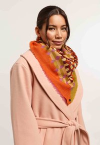 Sciarpa colorata con motivo geometrico in tonalità arancione, rosa e verde, realizzata in tessuto morbido con bordi sfrangiati, indossata sopra un cappotto rosa pallido.