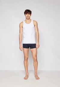 Maiou alb din bumbac și pantaloni scurți tip boxer bleumarin pe un model masculin, stând desculț în fața unui fundal simplu.