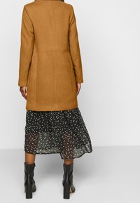 Brown wool-blend coat with a tailored fit and long sleeves, paired with a sheer black skirt featuring a small floral pattern and ankle boots.