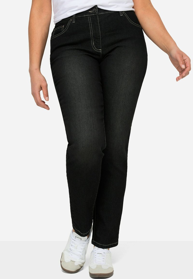 Jeans in denim nero con cuciture gialle a contrasto e taglio dritto. Indossati con sneakers bianche. Design minimalista, stile classico.
