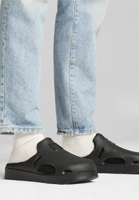 Crne slip-on cipele s teksturiranom površinom, ventilacijskim rupama i podstavljenim unutrašnjim dijelom, uparene s svijetloplavim farmerkama s manžetama i bijelim čarapama.