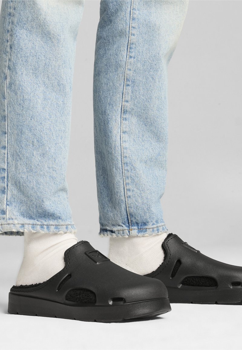 Crne slip-on cipele s teksturiranom površinom, ventilacijskim rupama i podstavljenim unutrašnjim dijelom, uparene s svijetloplavim farmerkama s manžetama i bijelim čarapama.