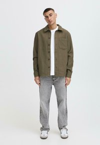 Olive green button-up shirt with a textured surface, a chest pocket, and black buttons, paired with faded grey jeans and white sneakers.
