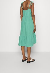 Robe en lin verte avec bretelles à nouer et ourlet à volants. Présente un corsage ajusté et une partie inférieure évasée. Portée avec des sandales noires et blanches.