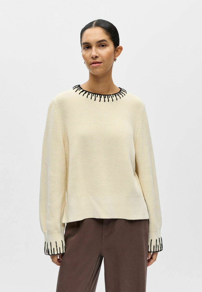 Femme aux cheveux foncés en chignon bas portant un pull en laine crème avec bordure noire au col et aux poignets, associé à un pantalon marron ample.