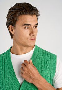 Grön stickad väst med flätmönster, med djup V-ringning och ribbad kant. Bärs över en vit T-shirt och visar en strukturerad yta.