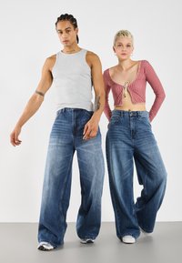 Graues Tanktop, kariertes rotes Cropped-Cardigan und weit geschnittene blaue Jeans. Auffällige Details sind eine Schleife am Cardigan und verschiedene Denim-Strukturen.