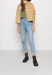 Giacca di velluto a coste color corda, t-shirt verde, jeans azzurri con risvolti leggeri, e stivaletti neri alla caviglia. Caratterizzata da due tasche frontali sulla giacca.