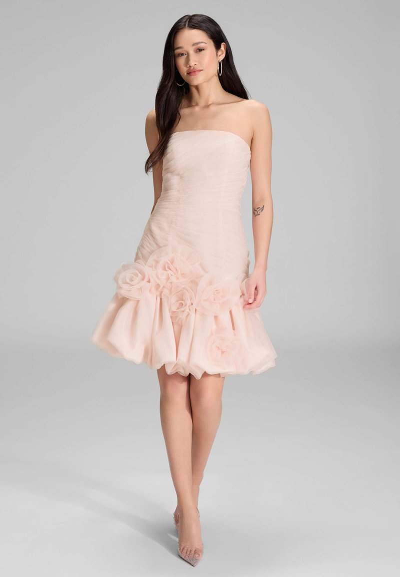 Robe rose clair sans bretelles avec un corsage froncé, jupe en tulle superposée et grandes appliques florales au bas. Talons transparents et pointus.