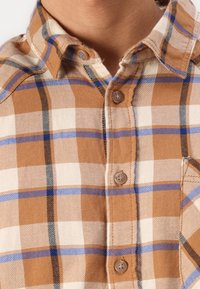 Close-up van een persoon in een bruine, witte, zwarte en blauwe geruite button-up shirt met een kraag en borstzak.