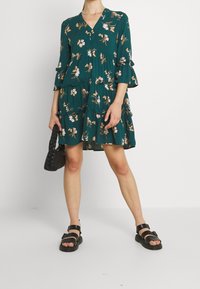 Turkos blommig klänning med V-ringning, trekvartslånga klockärmar, vågiga lager och en lös passform; kombinerad med svarta remsandaler och en handväska.