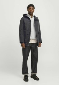 Marine vandtæt jakke med hætte, frontknaplukning, to store lommer, lagdelt over en lysegrå sweater, parret med mørke denim jeans og sorte sko.