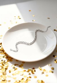Silverfärgad kedjearmband på en vit tallrik, med ovala länkar, justerbar längd, med spridda guldkonfetti i bakgrunden.