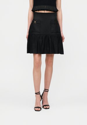 Kobieta ubrana w czarną mini spódniczkę z przednimi kieszeniami i złotymi klamrami, w połączeniu z czarnymi sandałami na wysokim obcasie z paskami.