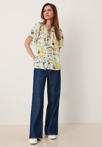 Bluse mit floralem Muster, kurzen Ärmeln und überwiegend gelber, blauer und schwarzer Farbgebung, mit einem Schlitz vorne und lockerer Passform, kombiniert mit dunklen, weiten Jeans.